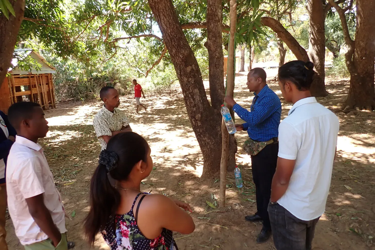 Community members and educators learning practical conservation tools A group of adults stand in a shaded forest area, observing a demonstration involving a water bottle and bamboo stick.