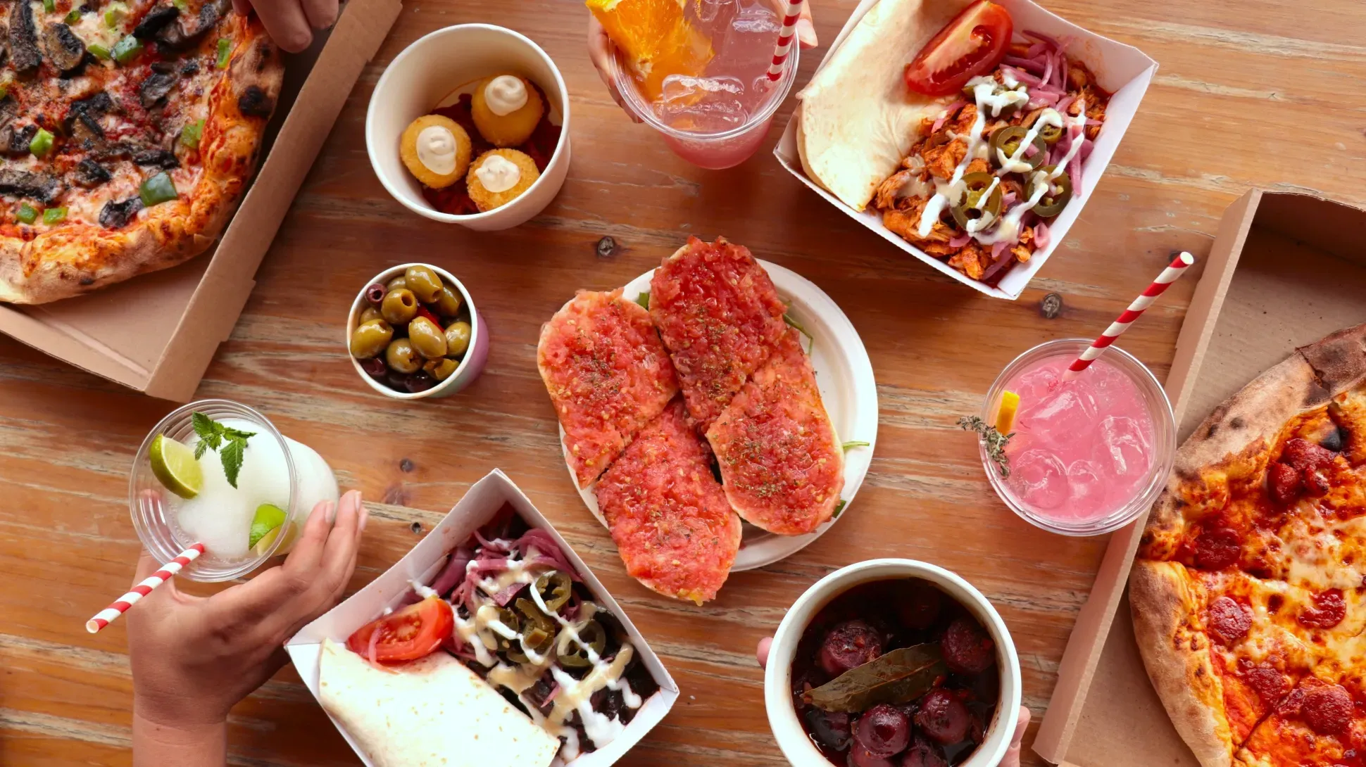 Food Village 2025 Top-down view of a table with pizza, shawarma wraps, bruschetta, olives, fried bites, sausages, and colourful drinks with straws.