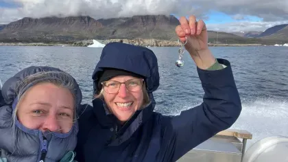 Two women wrapped up warm on a boat. One is holding a tiny disco ball as the mountains of Disko Island are visible in the distance