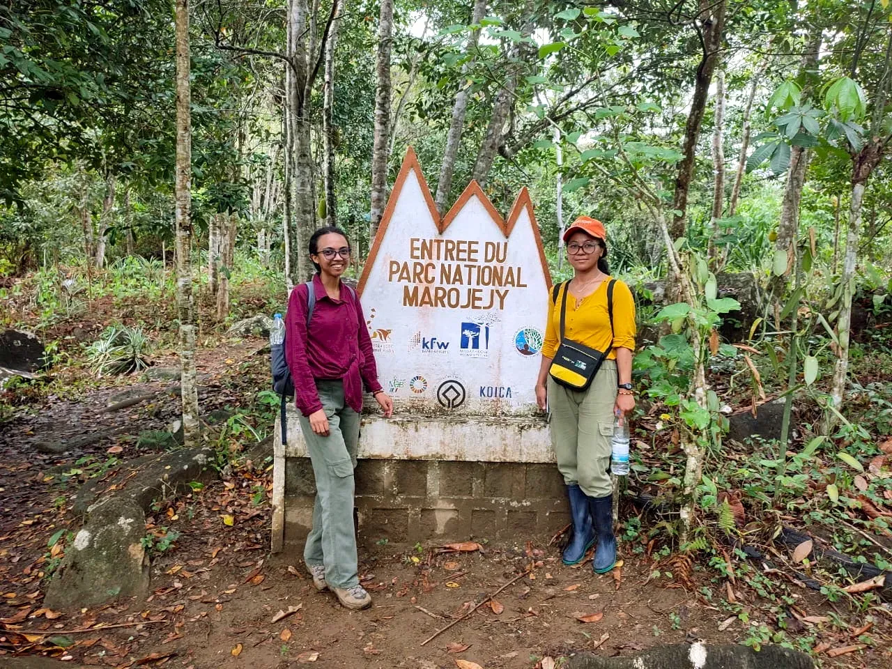 Two of Lova's students - Lolie (left) and Onja (right) - on a fieldtrip to Marojejy National Park Two people standing either side of a large wooden sign in a forest