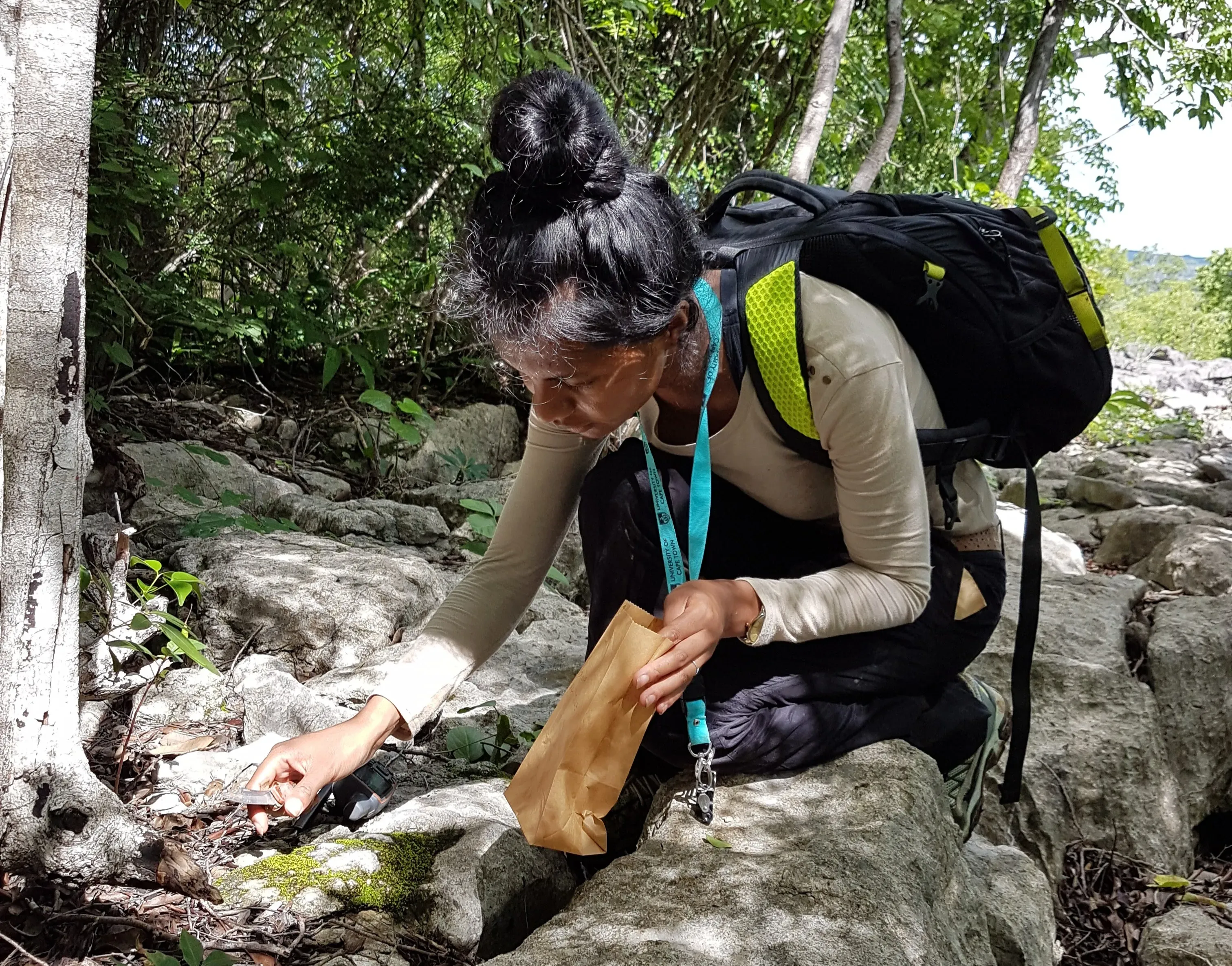 Lova collecting bryophytes in Ankarana national park, Madagascar. © Lova Marline Lova crouched down among some rocks with a small knife in one hand and a paper bag in the other